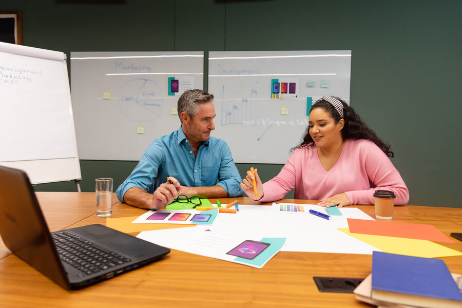 Estrategias para atraer talento humano implementacion - Dos personas en reunión de trabajo creativo, revisando paletas de colores y diseños en una sala con pizarras y gráficos.