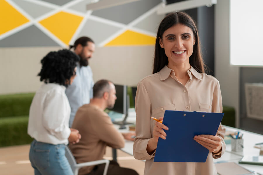 Técnicas de selección de personal conclusion - Mujer sonriente con portapapeles en oficina moderna con equipo de trabajo al fondo.